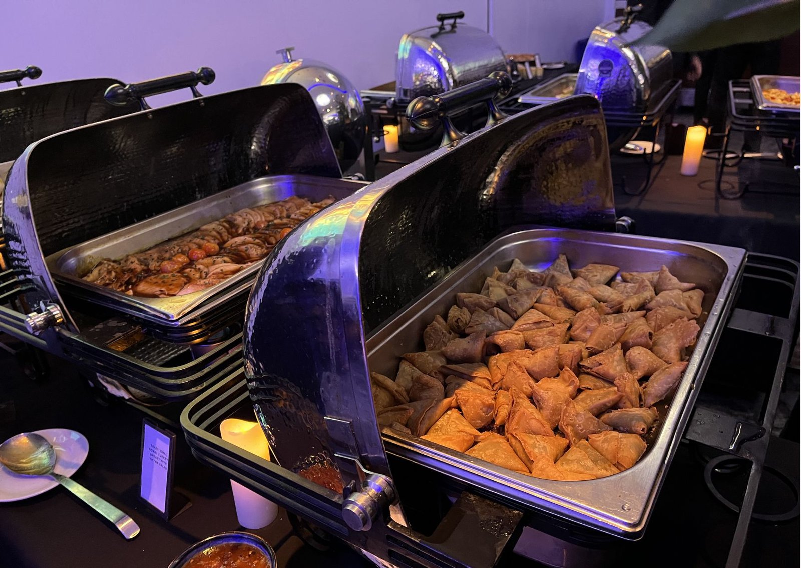 Buffet chafing dishes with warm appetizers, including triangular samosas in the front tray, at a dimly lit event hall.