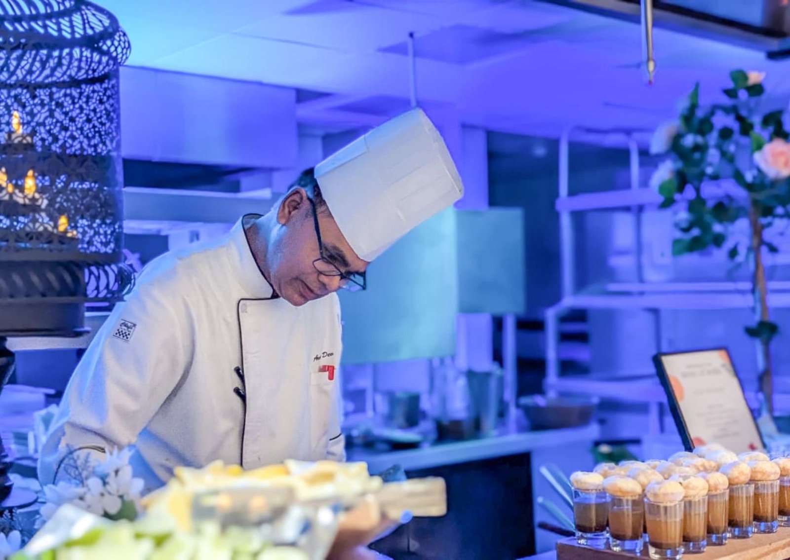 Chef in a white uniform and tall hat bending over a pastry station in a blue-lit kitchen, with desserts on display