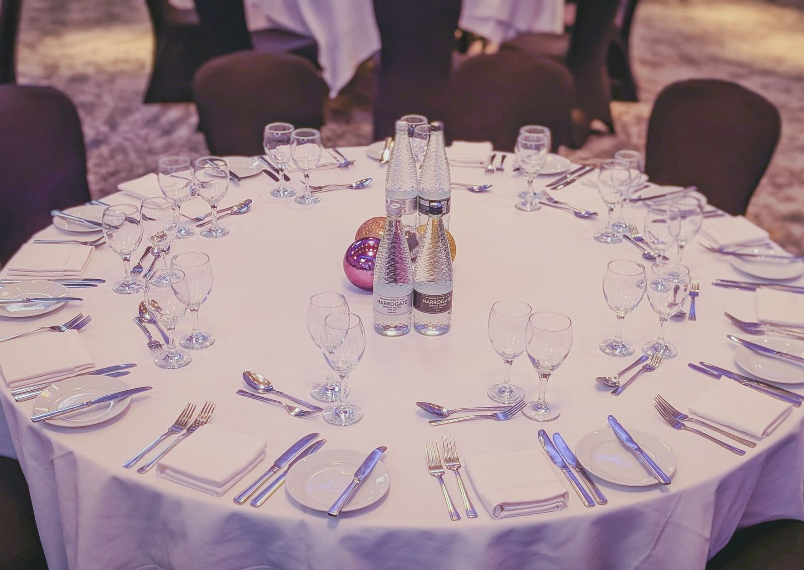 Round banquet table dressed in white with full place settings, glassware, and center bottles of water, framed by empty chairs.