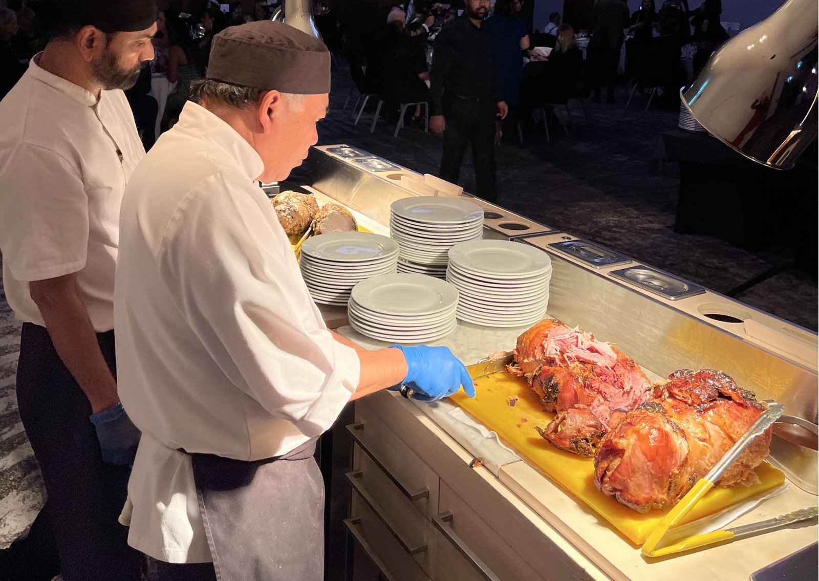 Two chefs in white uniforms carve roasted meat at a buffet with stacked plates nearby.