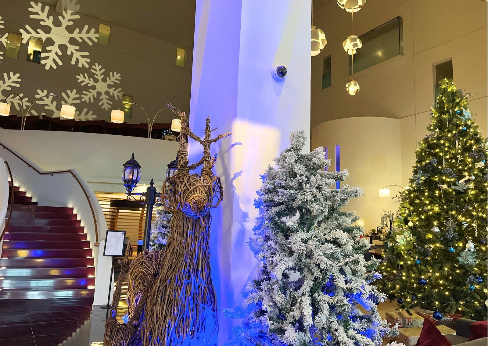 Lobby with Christmas decorations: snow-dusted trees, warm lights, and a large wicker reindeer sculpture beside a curved red-carpet staircase.