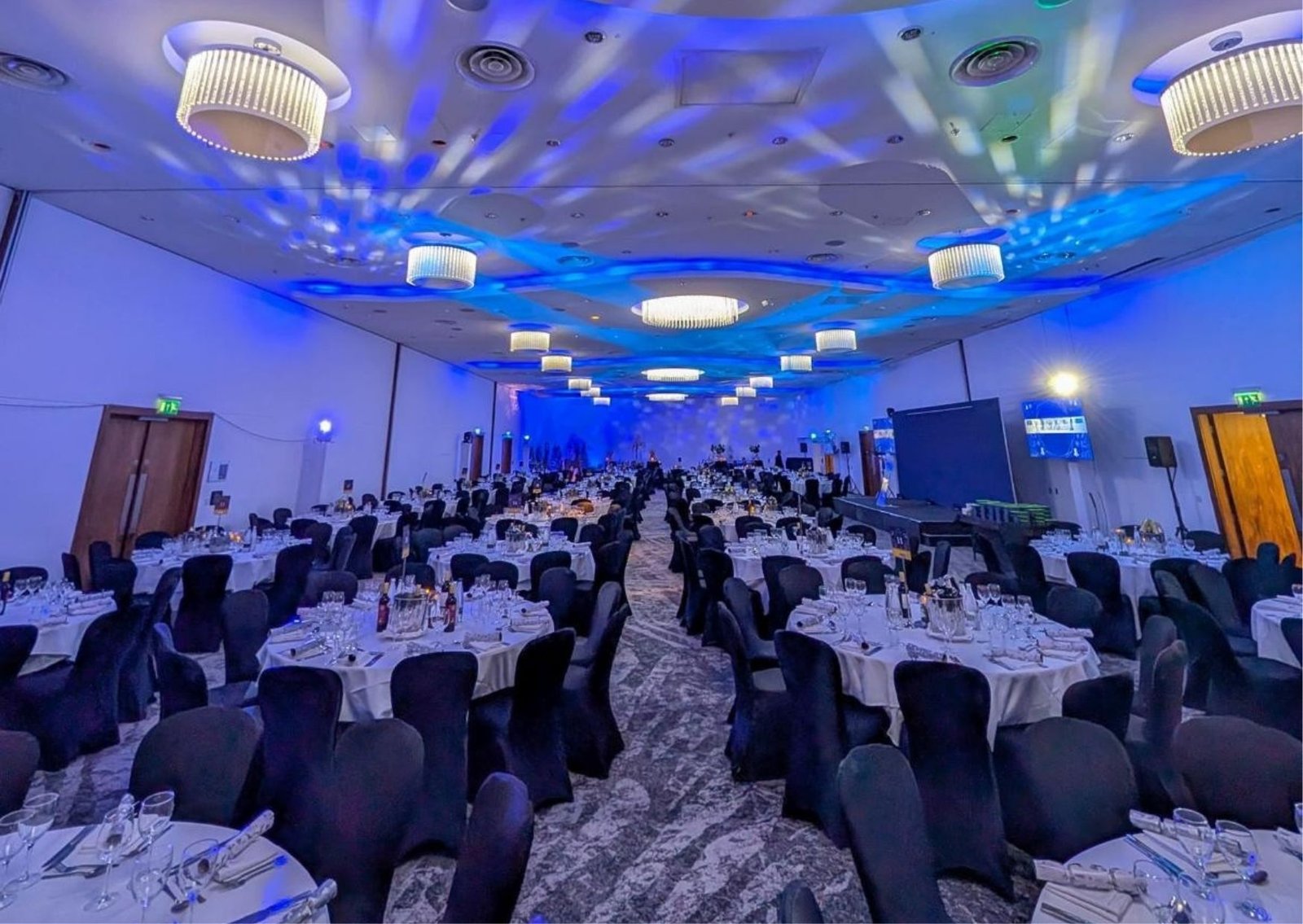 Banquet hall set for a formal dinner with round tables, black chair covers, white tablecloths, and blue stage lighting.