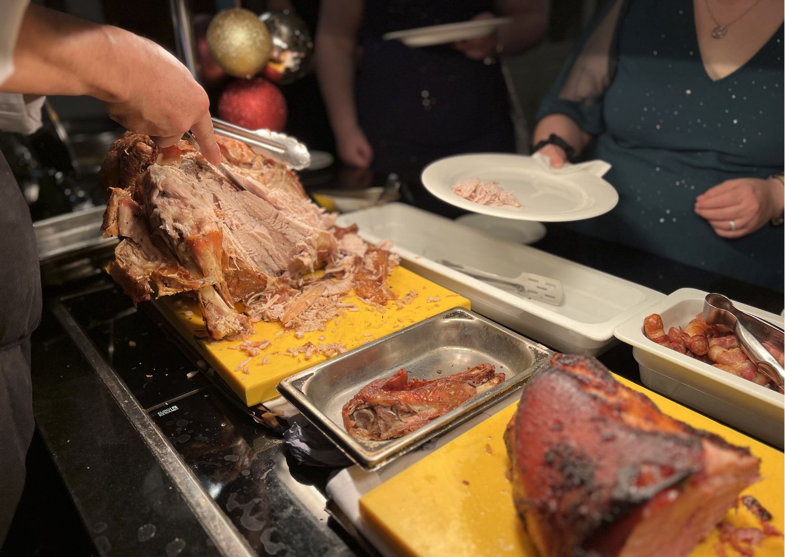 A cook carves a large roasted turkey on a yellow carving board, shredding meat as a guest holds a plate nearby.