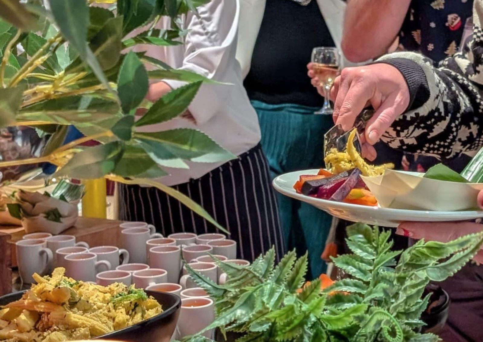 Hands serving food at a festive buffet with plates, cups, and greenery decorations. (informative)