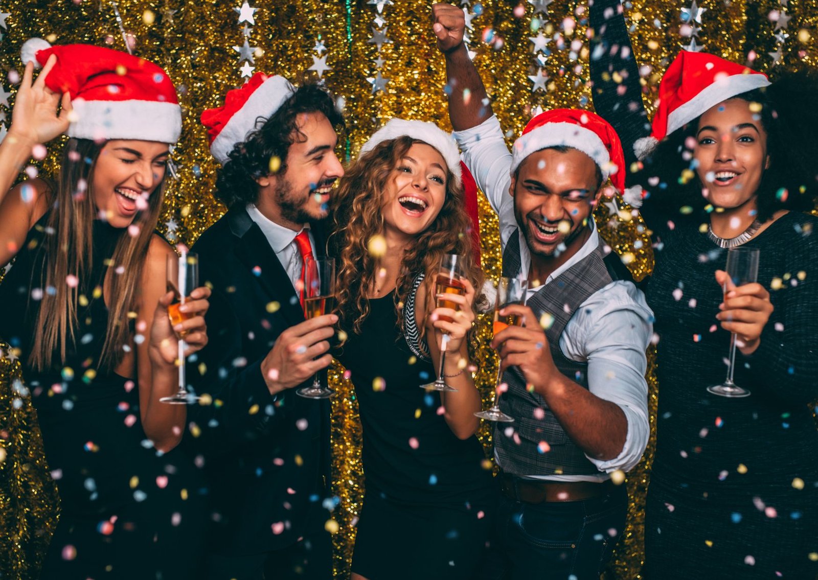 Group of six friends in Santa hats toasting with champagne, surrounded by confetti and a gold backdrop.