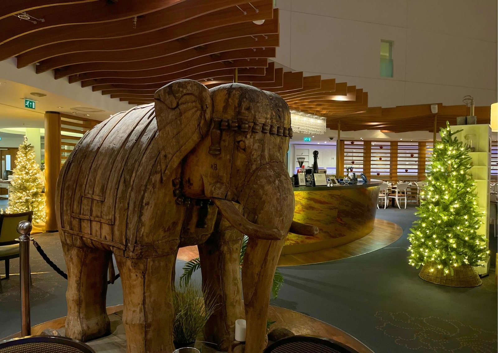 Large carved wooden elephant sculpture indoors near a bar, with Christmas trees and festive lights in the background.