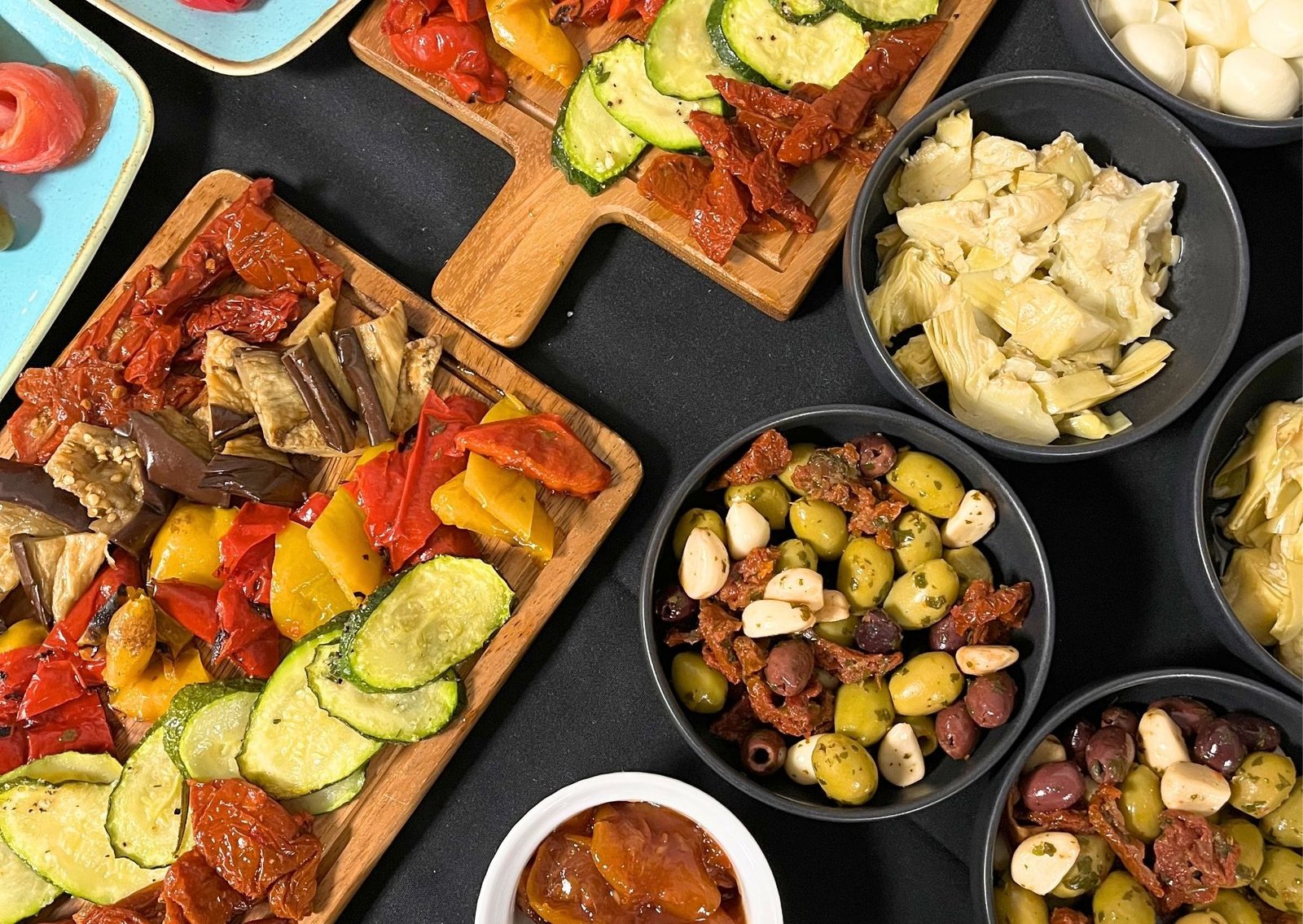 Assorted Mediterranean tapas: roasted zucchini, sun-dried tomatoes, grilled peppers on wooden boards with bowls of olives and artichokes nearby.