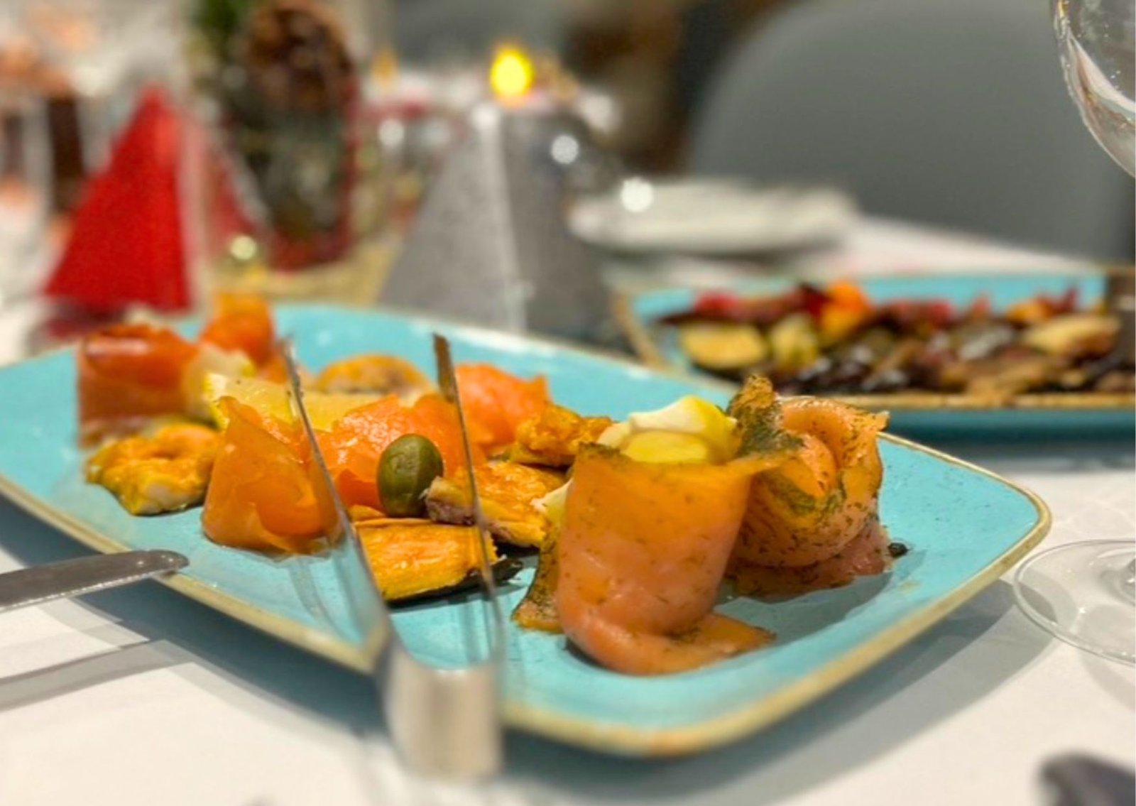 Turquoise plate with smoked salmon, orange vegetables, olives, and herbs on a dining table (blurred background).