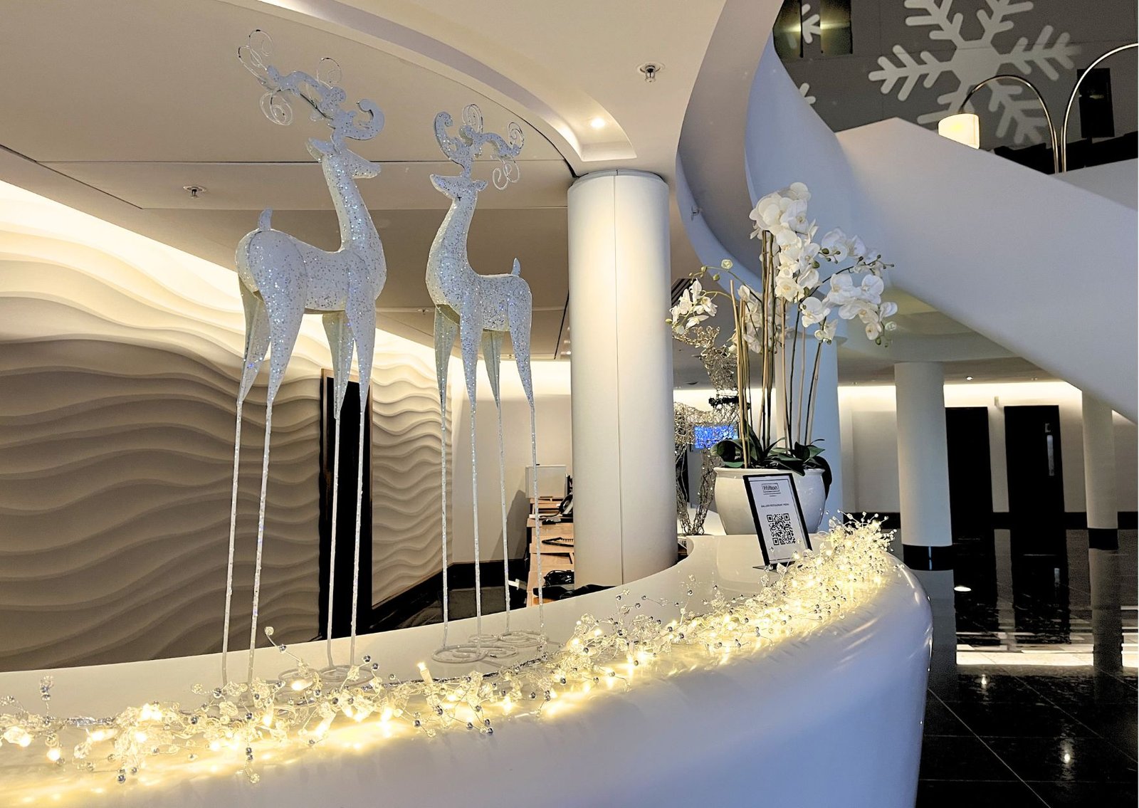 Ice-white glitter reindeer sculptures on a curved white reception desk with warm string lights in a modern hotel lobby.