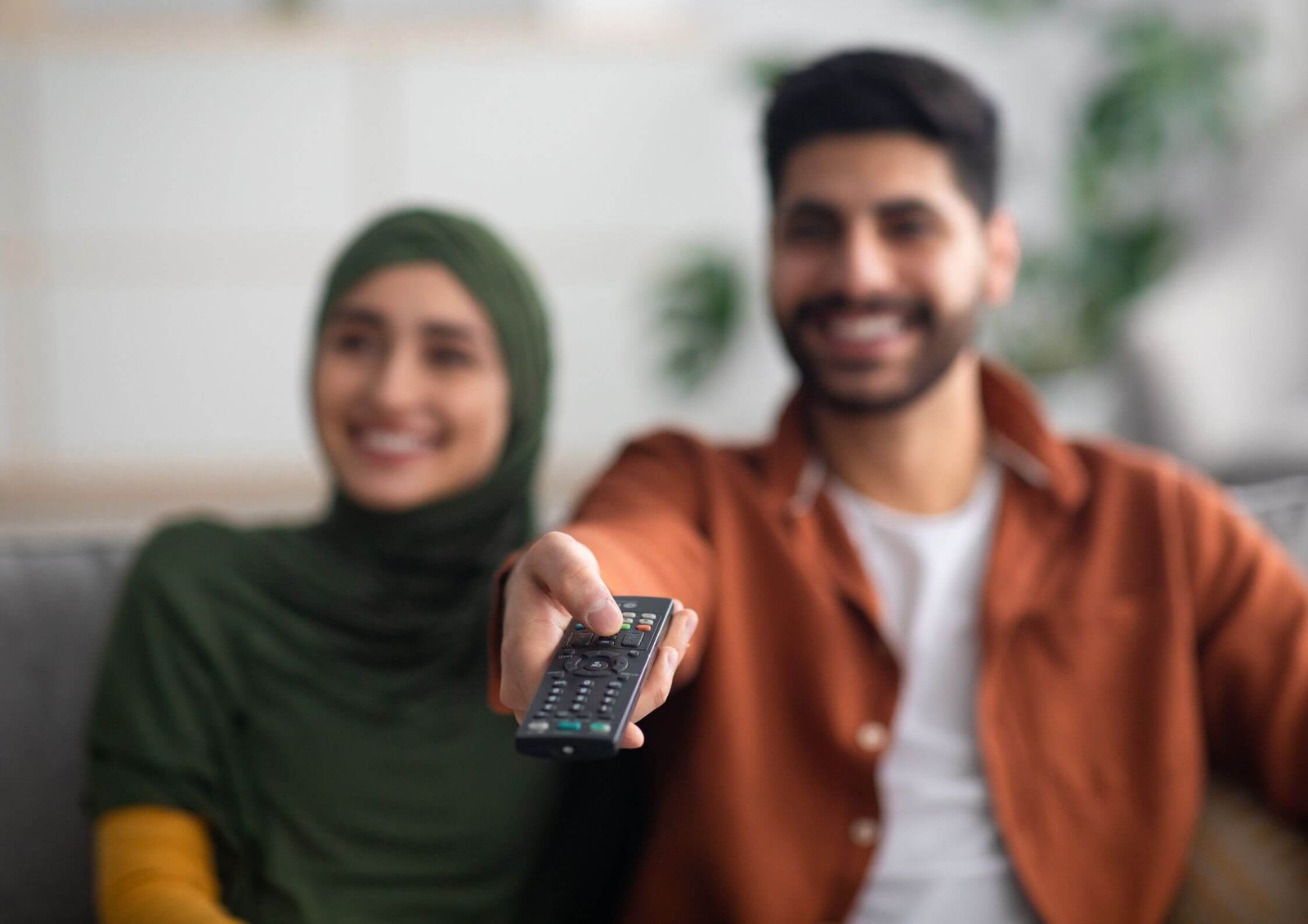 Two friends smiling for a selfie; a remote control is extended toward the camera in the foreground.