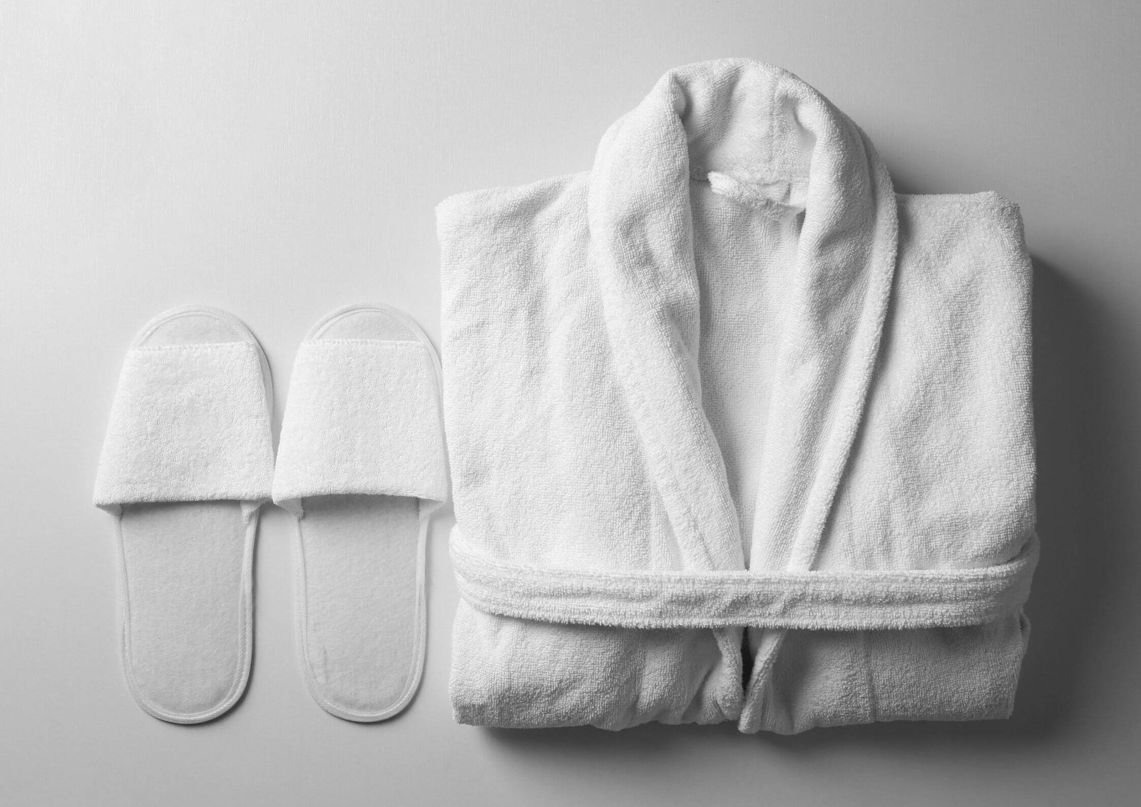 White towel-wrapped bathrobe laid over a surface with matching white slippers beside it, shown in grayscale for spa-style display.