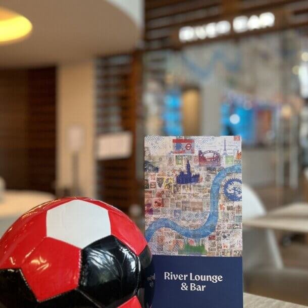 Red-and-black soccer ball in foreground with a travel-stamp collage brochure and a River Lounge & Bar sign in a blurred lounge interior