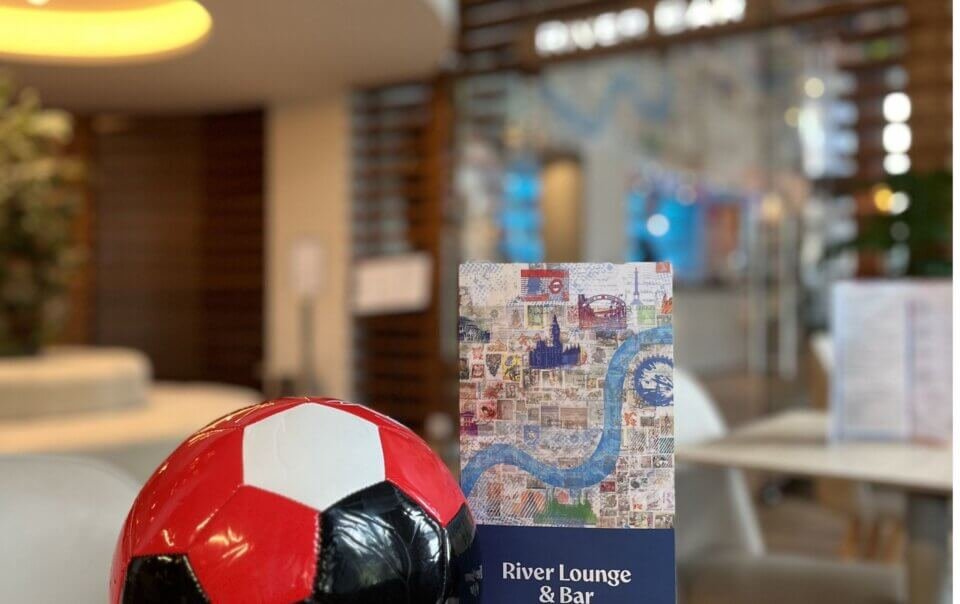 Red-and-black soccer ball in foreground with a travel-stamp collage brochure and a River Lounge & Bar sign in a blurred lounge interior