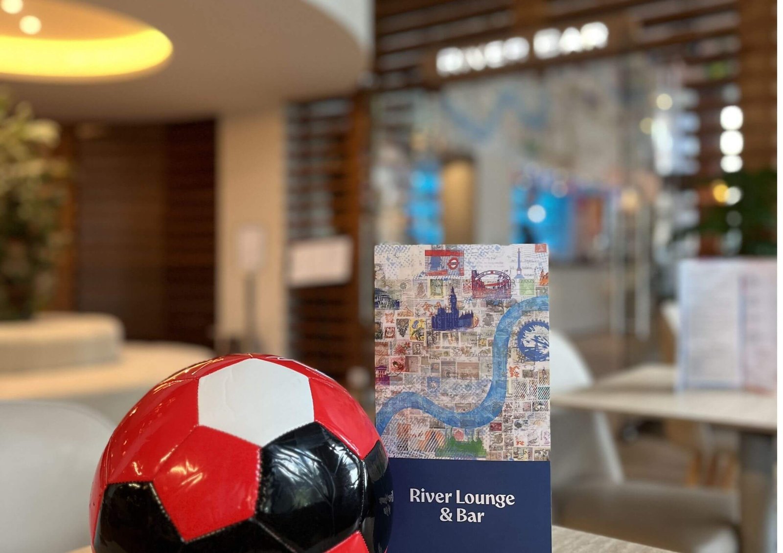 Red-and-black soccer ball in foreground with a travel-stamp collage brochure and a River Lounge & Bar sign in a blurred lounge interior