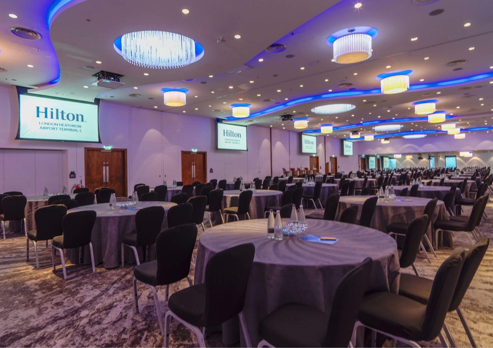 Banquet hall set up with round tables, black chairs, and Hilton-branded screens along the wall.