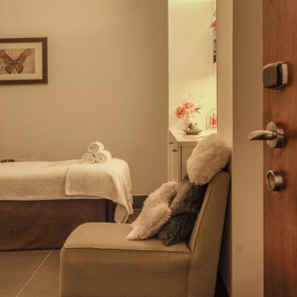 Spa treatment room featuring a massage bed with white towels, a fluffy chair, and warm beige walls with butterfly artwork.