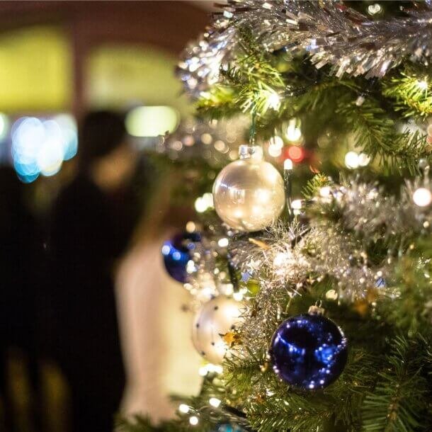 Close-up of a Christmas tree with blue and clear baubles and silver tinsel, soft party lights blurred in the background.