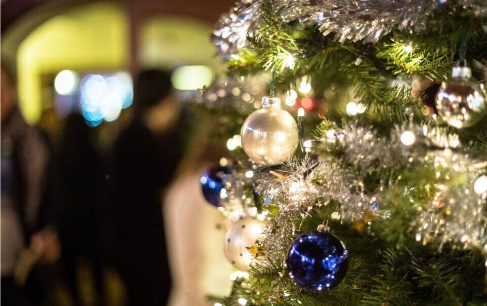 Close-up of a Christmas tree with blue and clear baubles and silver tinsel, soft party lights blurred in the background.