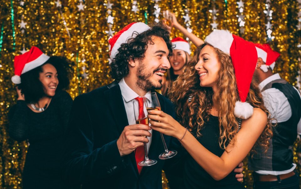 Group of friends in Santa hats toasting with champagne at a holiday party against a gold glitter backdrop in festive mood.