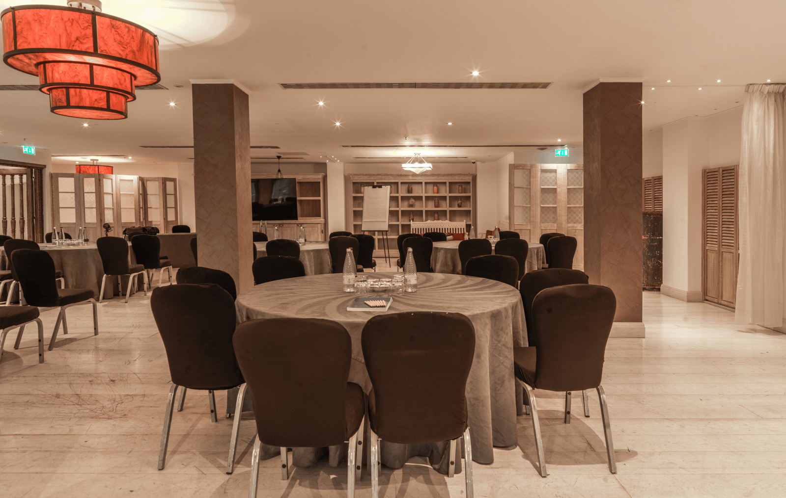Banquet hall with round tables covered in gray cloth and black chairs, warm lighting, and shelves in the back, ready for a conference or reception.