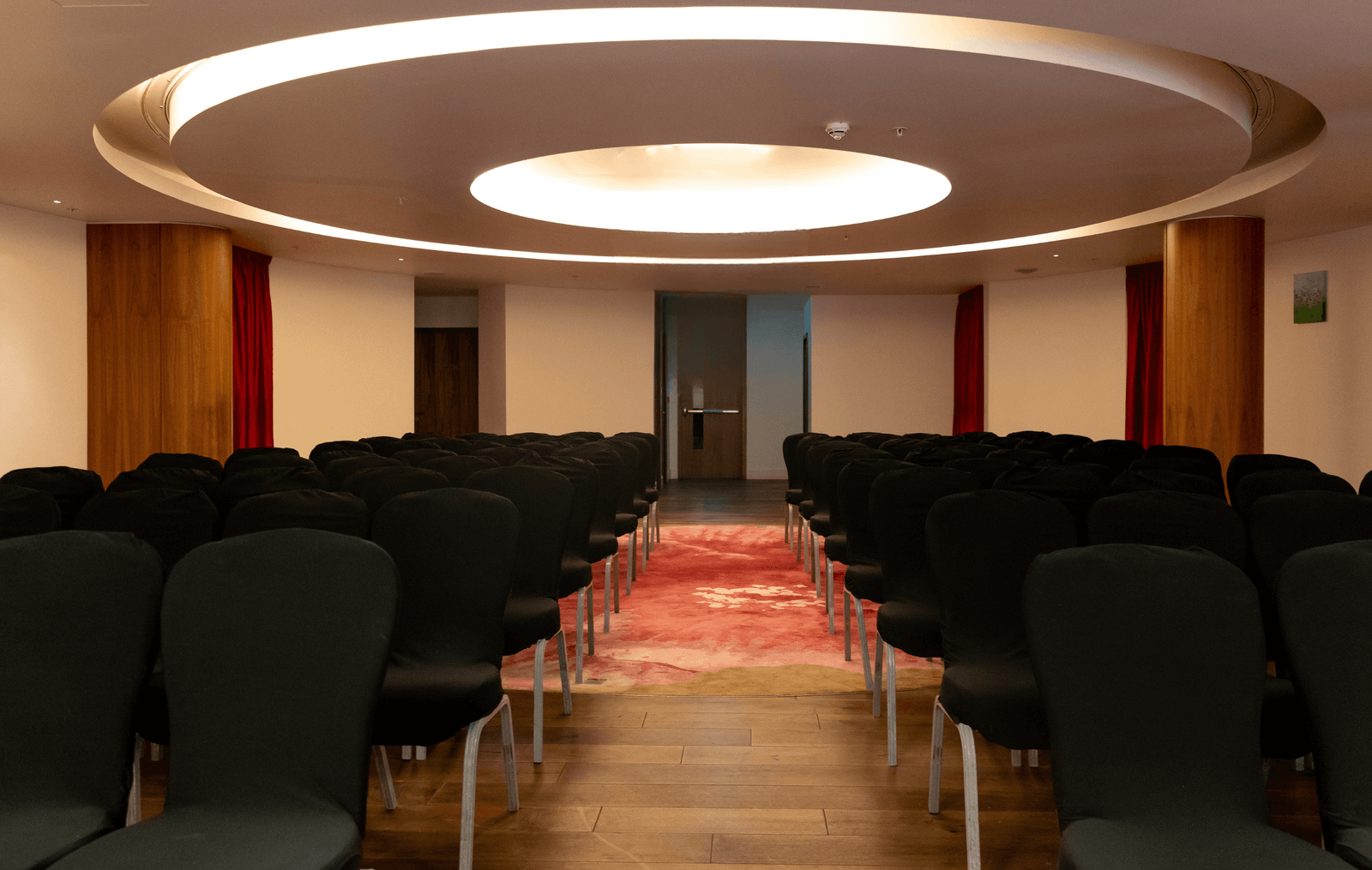 Auditorium with black chairs arranged in rows, facing a back doorway, under a circular illuminated ceiling fixture and red carpet.