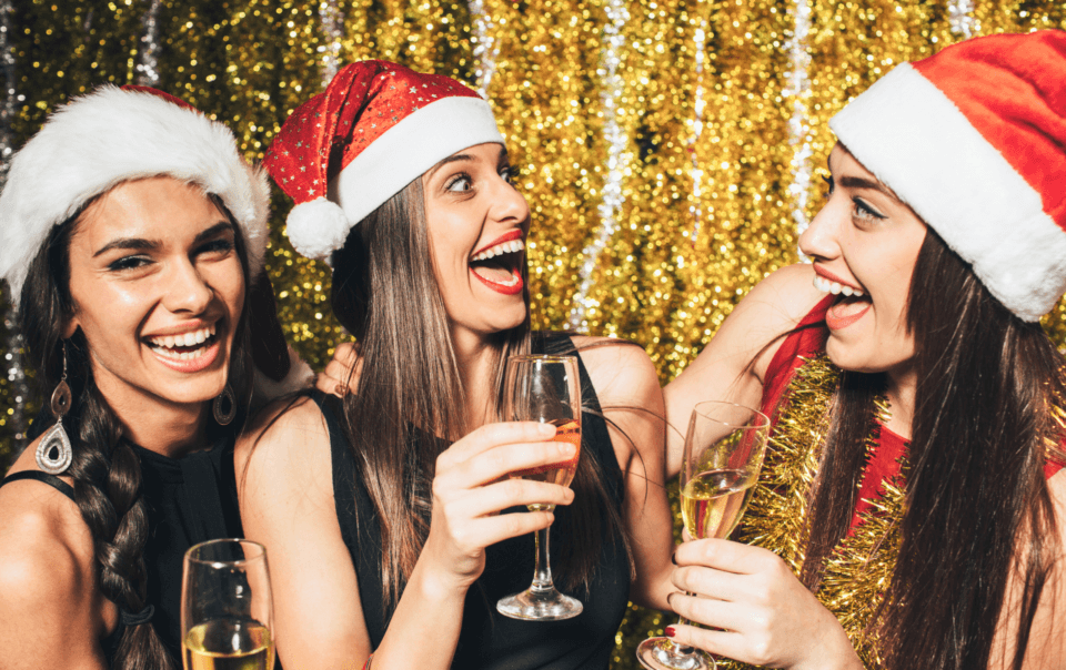 Three friends in Santa hats toasting with champagne at a festive party, gold glitter backdrop behind them.